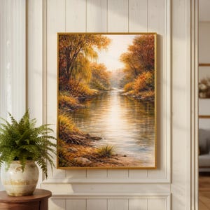 May include: A framed landscape painting depicts a serene river scene in autumn. The artwork features a golden frame and showcases a river flowing through a forest with trees in shades of orange, yellow, and brown. The water reflects the warm colors of the foliage.