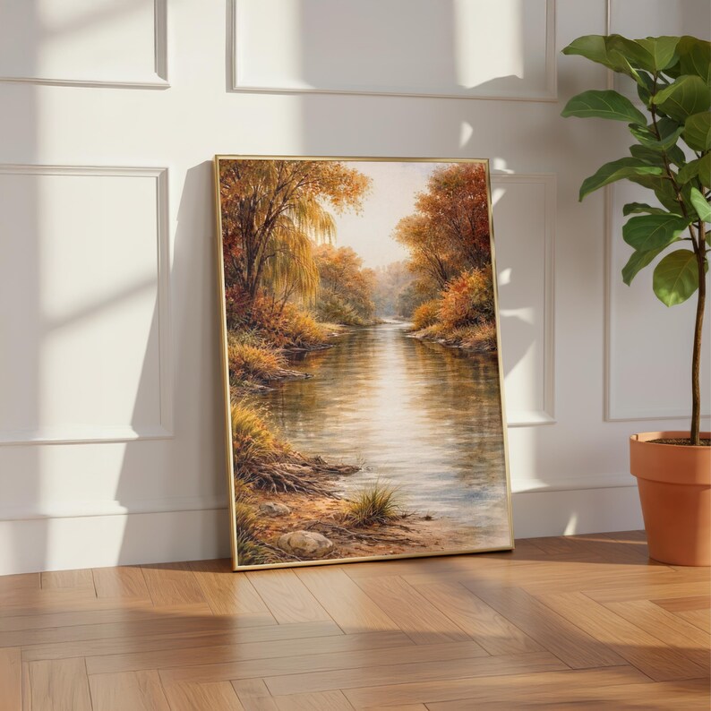 May include: A framed landscape painting depicts a serene river scene. The artwork features a golden frame and showcases a river flowing through a forest with autumn foliage in shades of orange, yellow, and brown. The painting is leaning against a white wall.