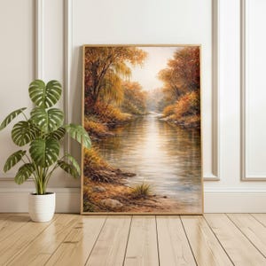 May include: A framed landscape painting depicts a serene river scene in autumn. The artwork features a golden frame and showcases a river flowing through a forest with trees in shades of orange, yellow, and brown. The water reflects the warm colors of the foliage.