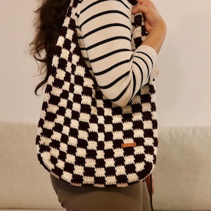Può includere: Una borsa hobo all'uncinetto con un motivo a scacchi marrone scuro e crema. La borsa presenta una struttura morbida e cadente con un'ampia tracolla e una piccola etichetta rettangolare in similpelle marrone sulla parte anteriore inferiore.