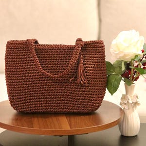 Può includere: Una borsa tote marrone all'uncinetto con una forma rettangolare e un manico intrecciato. La borsa presenta un nappa decorativa ed è esposta su un tavolo di legno. È un articolo fatto a mano.