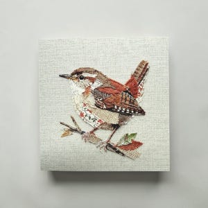 May include: A detailed textile art piece featuring a small bird perched on a branch. The bird has brown, red, and white patterned feathers. The artwork is mounted on a square, light gray fabric background.