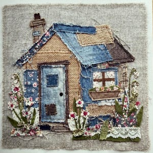 May include: A textile artwork showing a small house with a denim roof, burlap walls, and a blue door. Floral fabric is used for the windows and surrounding the house. The artwork is set against a textured, neutral-toned backdrop.