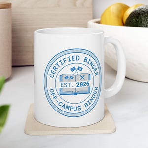 May include: White ceramic mug featuring a blue circular design with the text "CERTIFIED BINGER OFF-CAMPUS BINGER EST. 2026". The design includes an open book, flags, and an "X". The mug sits on a beige coaster.