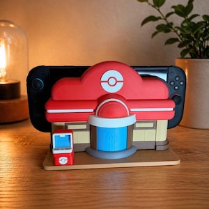 May include: A red, white, and tan Nintendo Switch dock shaped like a building with a red roof and a white and red logo. A small red and white accessory is on the left. The dock is on a wooden surface.