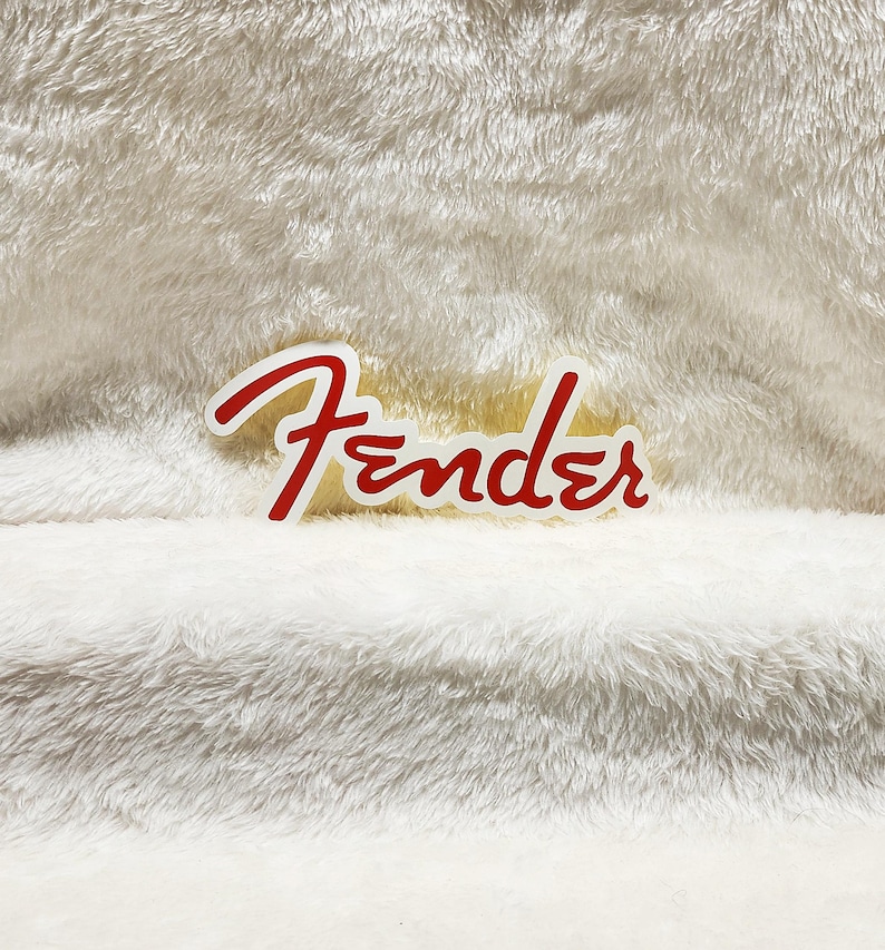 K&ouml;nnte beinhalten: Ein wei&szlig;-roter Fender-Logo-Aufkleber. Das Wort "Fender" ist in roter Schreibschrift mit wei&szlig;er Umrandung geschrieben. Der Aufkleber befindet sich auf einer wei&szlig;en, flauschigen Oberfl&auml;che.