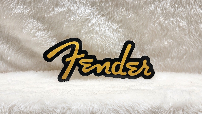 May include: A black and gold Fender logo sticker. The word "Fender" is written in a stylized gold font with a black outline. The sticker is on a white, textured background.