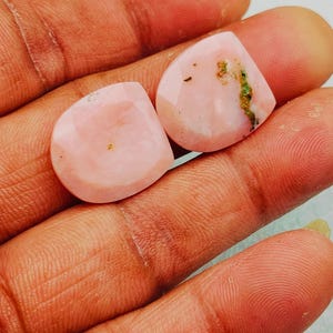 May include: Two pink gemstones with a faceted cut, displayed on a hand. The stones have a soft, translucent appearance with subtle variations in color. One stone has a small patch of green and brown inclusions.