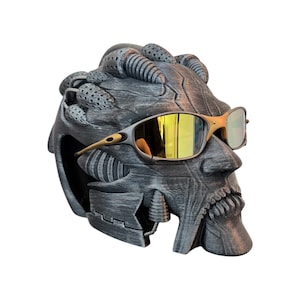 May include: A dark gray, textured, biomechanical-style head sculpture featuring intricate mechanical details and ridges. The sculpture is wearing a pair of metallic gold-toned sunglasses with reflective yellow lenses that cover the eye area.