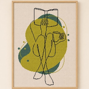 May include: A framed cross-stitch art piece featuring a minimalist line drawing of a person reading a book and holding a mug. The design is in black thread on a cream-colored background, with green and teal abstract shapes. The frame is made of light-colored wood.