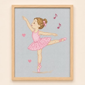 Peut inclure: Une broderie au point de croix représentant une ballerine en tutu et chaussons roses, avec des cheveux bruns en chignon ornés d'un nœud rose. Le motif inclut deux cœurs et deux notes de musique roses sur fond bleu clair, encadré en bois clair.