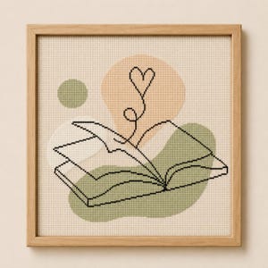 May include: A framed cross-stitch artwork depicting a black line drawing of an open book with a heart design above it. The background has abstract shapes in peach, green, and beige. The wooden frame provides a natural aesthetic.