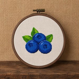 Puede incluir: Un bastidor de bordado circular de madera que contiene tela Aida blanca con un patrón de punto de cruz de tres arándanos azules vibrantes y tres hojas de color verde brillante centradas, asegurado con un tornillo de metal.