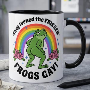 May include: White ceramic mug with a black interior and handle. The mug features a cartoon frog in front of a rainbow, with the text "They turned the FRICKEN' FROGS GAY!" in a bold, playful font. Pink flowers and blue stars are also included.