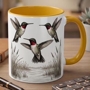 May include: White ceramic mug with a yellow interior and handle. The mug displays three illustrations of hummingbirds in flight, set against a marsh scene with reeds and water, creating a nature-themed design.