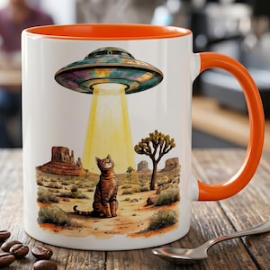 May include: White ceramic mug with an orange interior and handle. The mug features a colorful illustration of a UFO beaming a light onto a cat in a desert landscape. The cat is brown and orange with stripes.