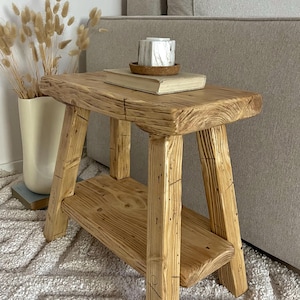 May include: A small rectangular wooden stool with a natural light brown finish, featuring a thick top surface, four sturdy legs, and a lower shelf. The wood grain is visible throughout the structure, showcasing a textured, rustic appearance.