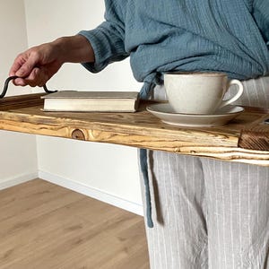 May include: A rectangular wooden serving tray featuring a natural wood grain finish with visible knots and dark, scorched edges. The tray includes two curved black metal handles attached to the sides, holding a white ceramic cup and saucer set.
