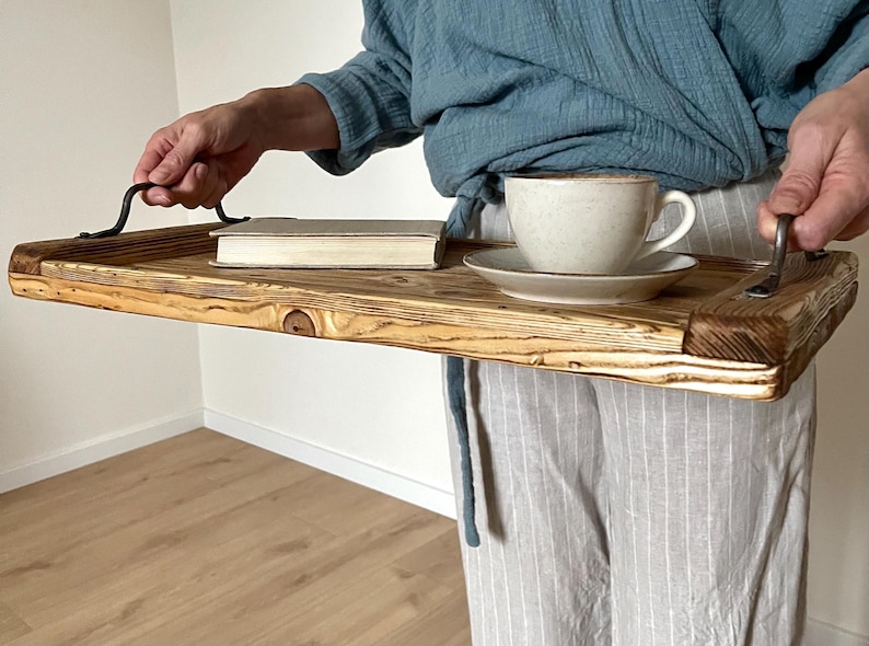 May include: A rectangular wooden serving tray featuring a natural wood grain finish with visible knots and dark, scorched edges. The tray includes two curved black metal handles attached to the sides, holding a white ceramic cup and saucer set.
