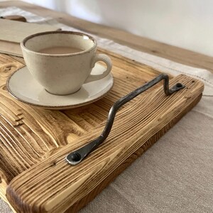 May include: A rectangular wooden serving tray featuring a prominent, dark, hand-forged metal handle secured with screws. The tray has a visible, textured wood grain finish. A small, speckled, light-colored ceramic cup and saucer containing a beverage sit on the tray.