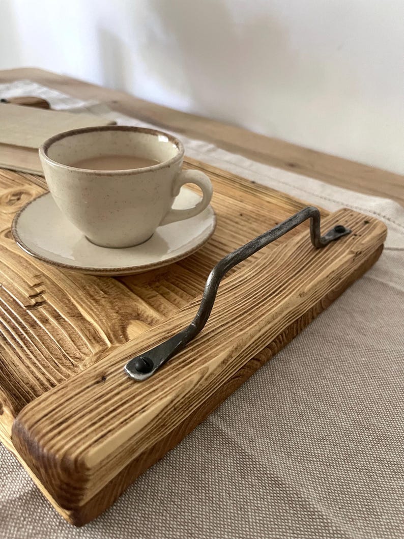 May include: A rectangular wooden serving tray featuring a prominent, dark, hand-forged metal handle secured with screws. The tray has a visible, textured wood grain finish. A small, speckled, light-colored ceramic cup and saucer containing a beverage sit on the tray.