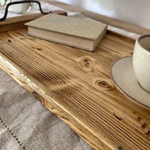 May include: A rectangular wooden serving tray featuring a natural light brown wood grain with visible knots and dark charred edges. The tray includes two black metal handles attached to the sides, providing a sturdy grip for carrying items.
