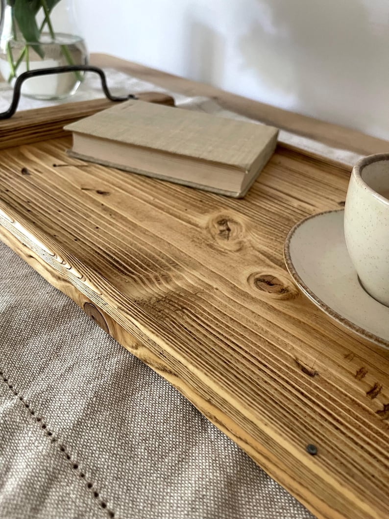 May include: A rectangular wooden serving tray featuring a natural light brown wood grain with visible knots and dark charred edges. The tray includes two black metal handles attached to the sides, providing a sturdy grip for carrying items.