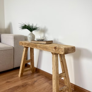 Op de afbeelding: Een rustieke houten bank met een natuurlijke afwerking. De bank heeft een rechthoekige bovenkant en A-vormige poten. Een kleine vaas met groen, een boek, een kaars en een kopje thee staan op de bank. De bank is ongeveer 122 cm lang.