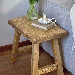 Peut inclure: Petite table d'appoint rustique en bois naturel. Le plateau rectangulaire repose sur quatre pieds robustes. Un vase en verre avec des tulipes roses, un livre et une bougie dans un bougeoir blanc sont posés dessus.