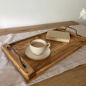 May include: A rectangular wooden serving tray with a scorched finish, featuring two dark metal handles on the sides. A beige ceramic cup and saucer containing a light brown beverage and a small closed book are placed on the tray surface.