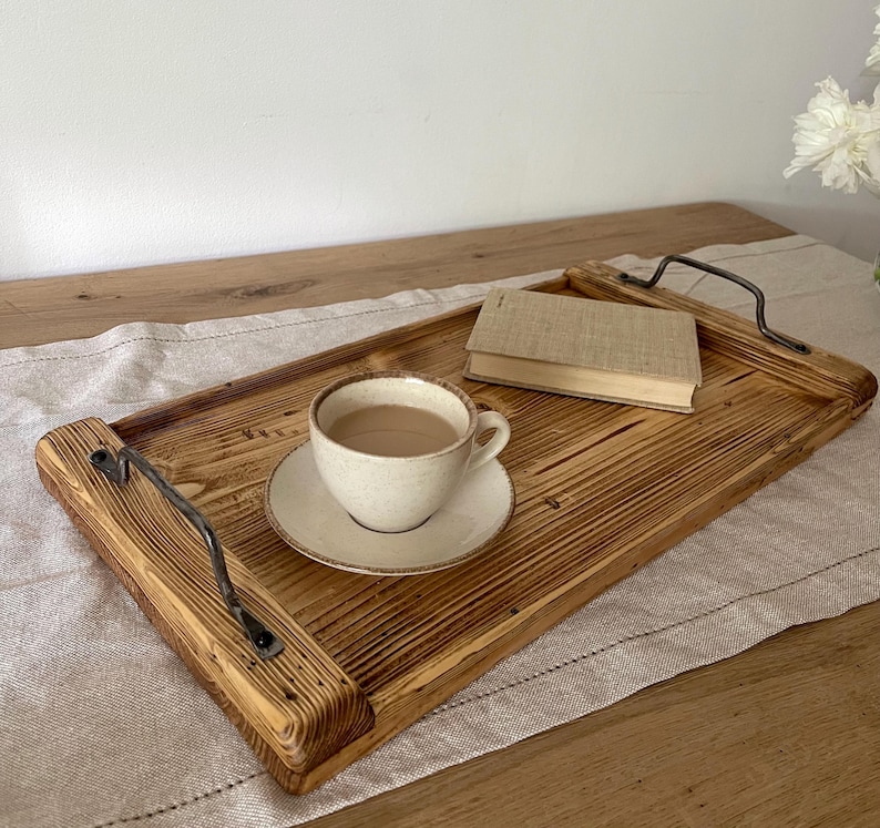 May include: A rectangular wooden serving tray with a scorched finish, featuring two dark metal handles on the sides. A beige ceramic cup and saucer containing a light brown beverage and a small closed book are placed on the tray surface.