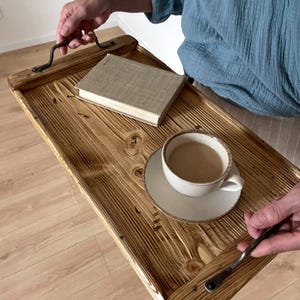 May include: A rectangular wooden serving tray with a dark, scorched wood grain finish and two black metal handles. A beige hardcover book and a ceramic cup filled with a light brown beverage on a matching saucer sit on the tray surface.