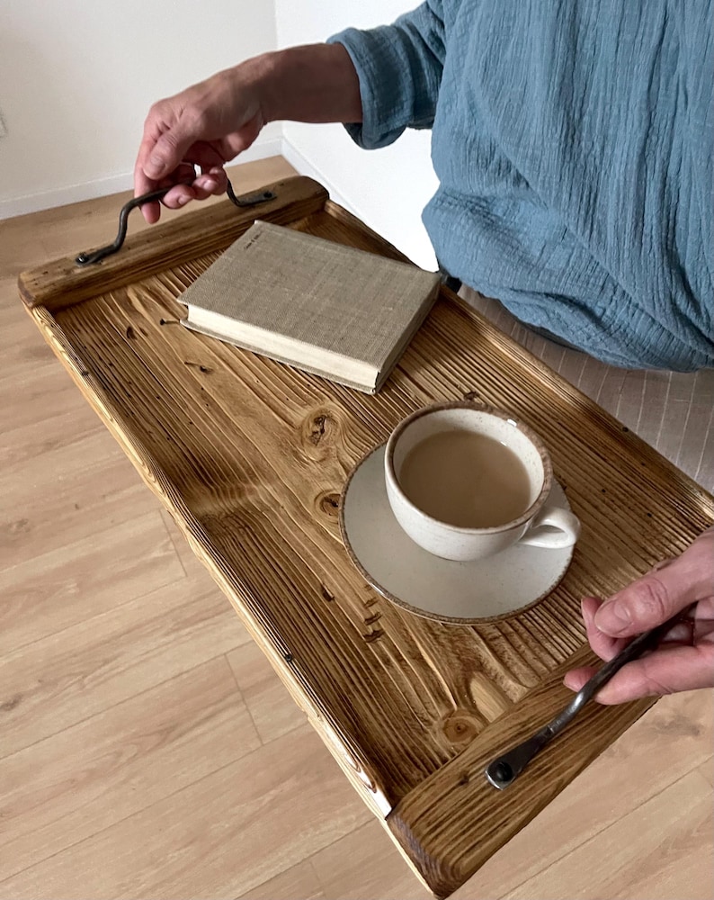 May include: A rectangular wooden serving tray with a dark, scorched wood grain finish and two black metal handles. A beige hardcover book and a ceramic cup filled with a light brown beverage on a matching saucer sit on the tray surface.