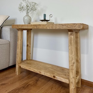 Può includere: Una consolle rustica in legno con un ripiano inferiore. Il tavolo è realizzato in legno chiaro con finitura naturale. Un piccolo vaso con fiori bianchi, libri e un portacandele con una candela turchese sono sopra. Il tavolo è lungo circa 100 cm.