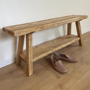 Può includere: Panca rustica in legno con ripiano inferiore, costruita in legno chiaro. La panca ha un aspetto naturale e non rifinito. Due paia di mocassini rosa sono posizionati sul pavimento davanti alla panca.
