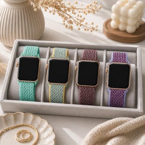 Puede incluir: Cuatro smartwatches con correas tejidas coloridas se exhiben en una bandeja gris. Las correas son en tonos turquesa, amarillo-verdoso, malva y morado. Las pantallas de los relojes son negras y las cajas son doradas.