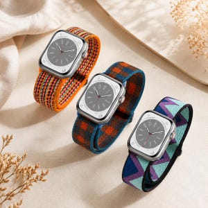 Könnte beinhalten: Drei Smartwatches mit farbenfrohen Armbändern. Eine hat ein orangefarbenes und mehrfarbiges gewebtes Band, eine andere ein blau-oranges Karomuster und die dritte ein blaues, lila und türkisfarbenes geometrisches Muster. Die Zifferblätter sind silberfarben mit grauen Ziffern.