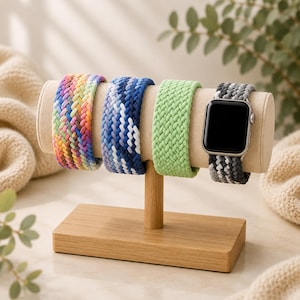 May include: A display of four braided watch bands in various colors. The bands are rainbow, blue with white accents, light green, and gray with white accents. They are displayed on a wooden stand with a beige cushion.