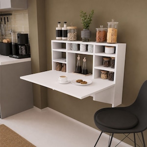 Peut inclure: Unité d'étagères de cuisine blanche murale avec une table rabattable. Les étagères contiennent des bocaux de nourriture, d'épices et d'objets décoratifs. Une tasse de café et des biscuits sont sur la table. Une chaise noire est au premier plan.