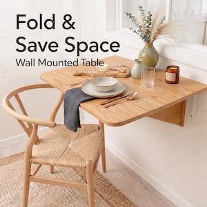 Peut inclure: Table murale en bois clair aux coins arrondis, avec un bol, une assiette et des couverts. La table est abaissée, avec une chaise en dessous. Le texte sur l'image indique "Fold & Save Space Wall Mounted Table."