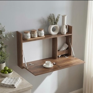 Peut inclure: Un bureau mural en bois à abattant avec une étagère au-dessus. Le bureau est ouvert, révélant une petite tasse et une soucoupe. L'étagère contient des pots, un vase avec de la verdure et un vase blanc. Le bureau mesure 76 cm de large.