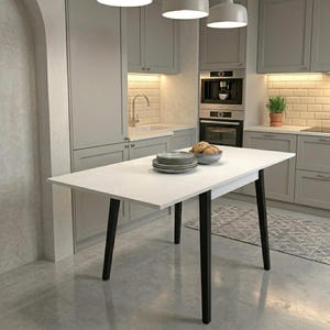 May include: A white kitchen table with black legs is the focal point of this image. The table is set with plates and a bowl of food. The kitchen features light gray cabinets, a built-in oven, and a small potted plant. The overall aesthetic is modern and minimalist.