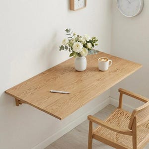 May include: A light oak wall-mounted desk with a white vase of flowers, a mug of coffee, and a pen. A wooden chair with a woven seat is positioned below the desk. A clock is mounted on the wall.