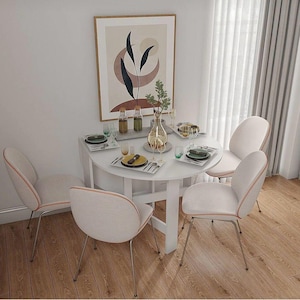 May include: A white, round dining table set for four with matching chairs. The chairs have light-coloured upholstery with orange trim and metal legs. A framed abstract art piece hangs on the wall behind the table. The table is set with plates, glasses, and a vase.