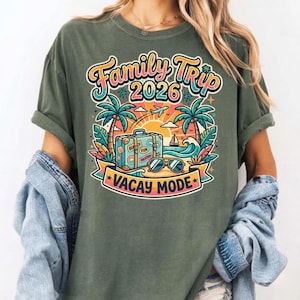 Può includere: T-shirt verde oliva con grafica colorata. Il design presenta "Family Trip 2026" sopra una scena di spiaggia con palme, una valigia, occhiali da sole e "Vacay Mode".