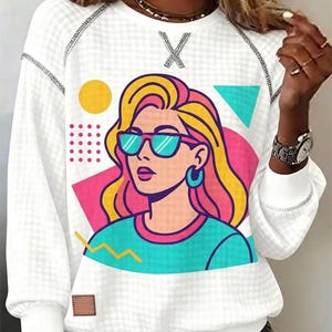 May include: White long-sleeved sweatshirt with a graphic of a woman wearing sunglasses. The graphic features a woman with yellow and pink hair, blue sunglasses, and a turquoise shirt. The sweatshirt has a textured pattern and a small brown patch.