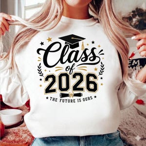 以下が含まれることがあります： 「Class of 2026」の文字と卒業帽子、星が描かれた黒と金のグラフィックが特徴の白いスウェットシャツ。「The Future is Ours」の文字も含まれています。卒業生へのプレゼントに最適です。
