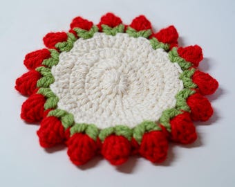 May include: A circular crochet coaster featuring a cream-colored center with a textured spiral stitch pattern. The outer edge is decorated with a border of small, three-dimensional red strawberry shapes accented with green yarn leaves.