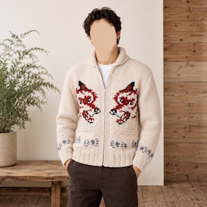 May include: A cream-colored knit zip-up cardigan featuring a shawl collar and a repeating red and black fox head pattern on the chest. The sleeves and hem are decorated with a small blue paw print motif, and the garment includes two front pockets.