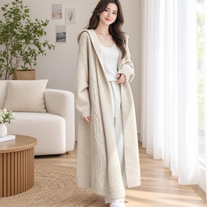May include: A long, beige, open-front hooded cardigan made of a knit fabric. The garment features long sleeves with ribbed cuffs, a hood, and a subtle vertical textured pattern running down the front panels. It is worn over a white tank top.